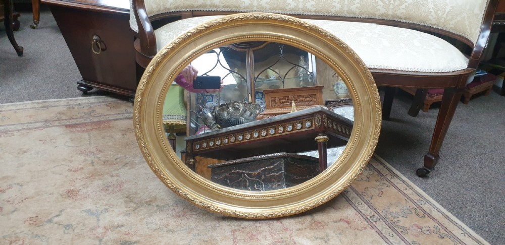 An Edwardian oval wall mirror featuring a gilt frame adorned with carved leaf and bead moulding. View V