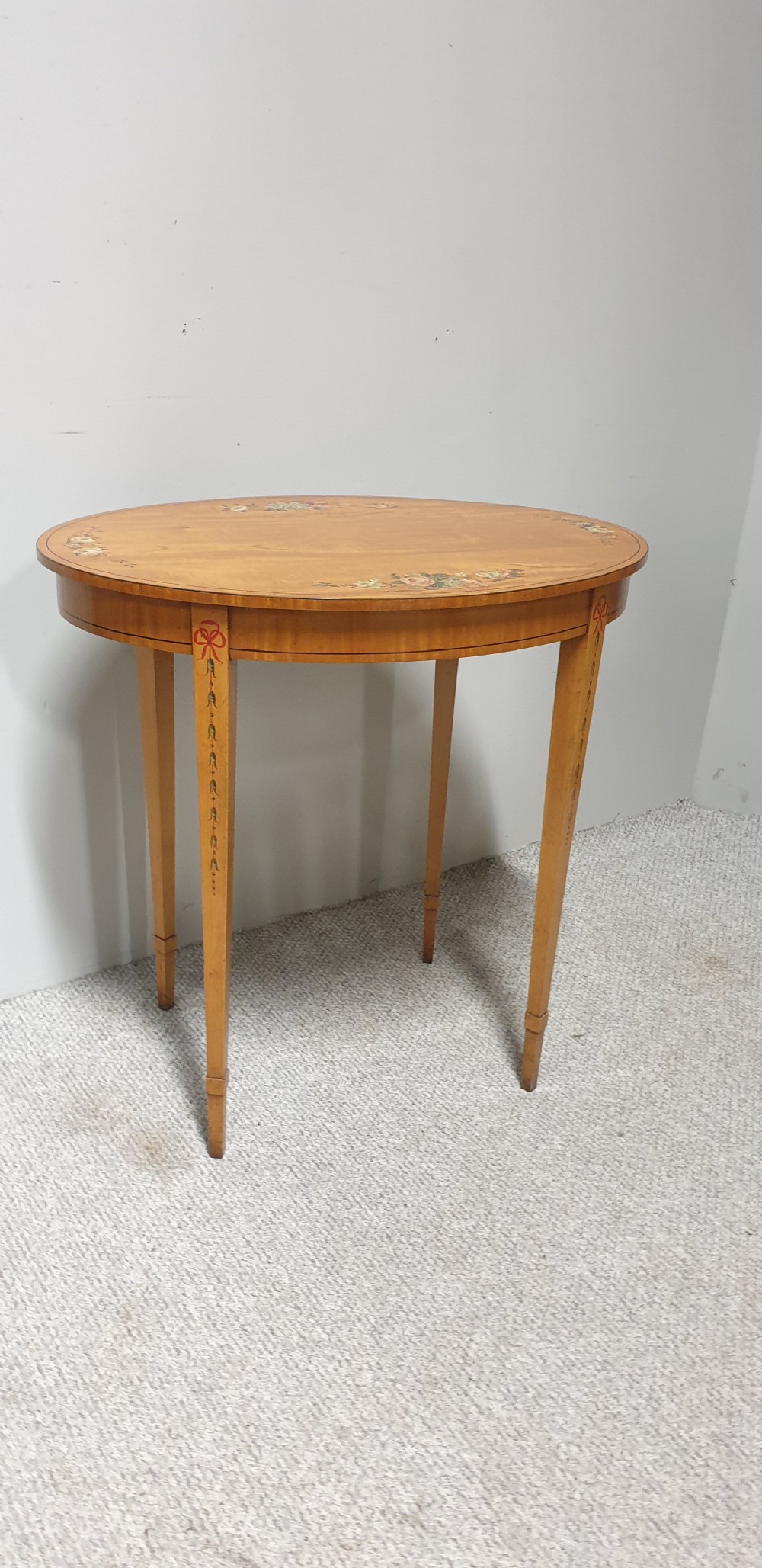 An oval satinwood occasional table featuring painted floral swags on the top surface and ribbon details along square tapered legs.