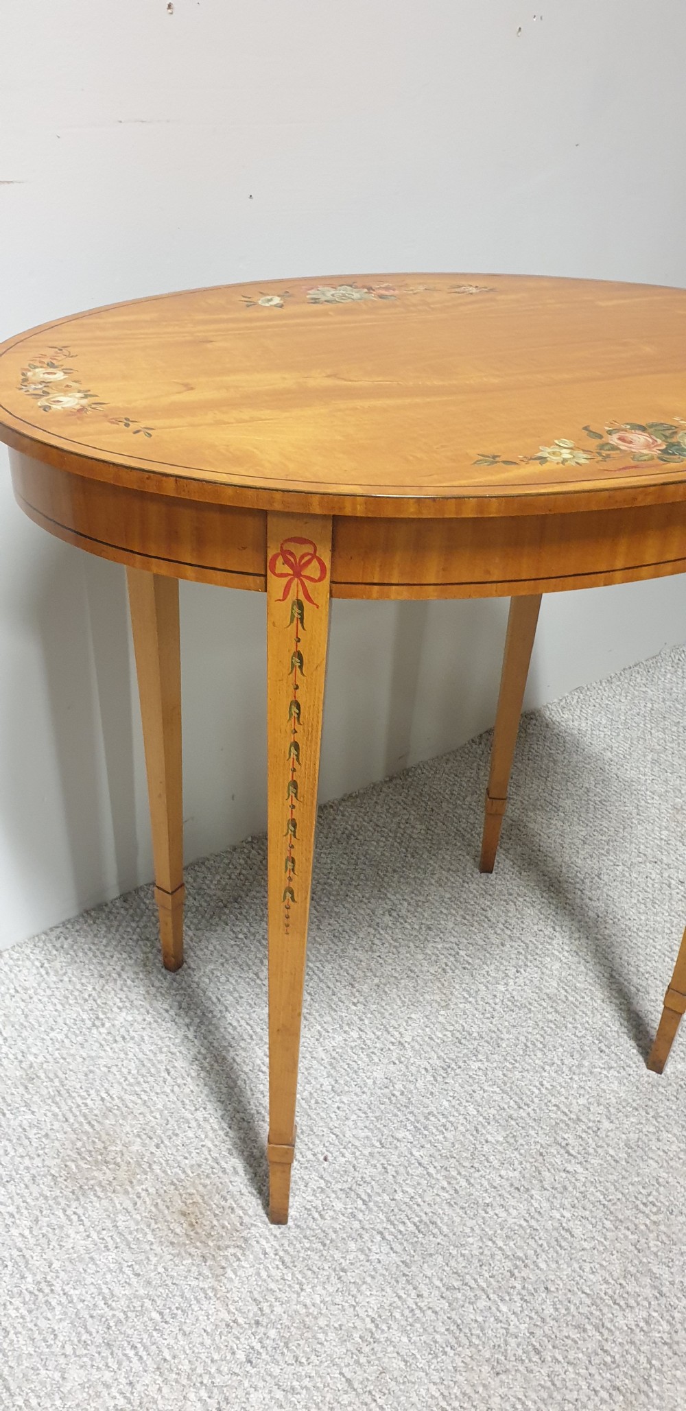 An oval satinwood occasional table featuring painted floral swags on the top surface and ribbon details along square tapered legs. View II