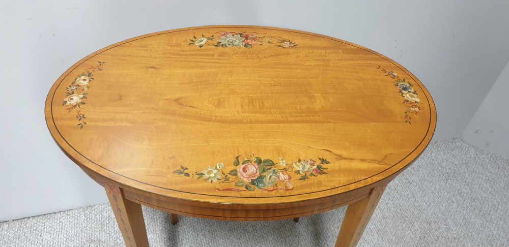 An oval satinwood occasional table featuring painted floral swags on the top surface and ribbon details along square tapered legs. View III