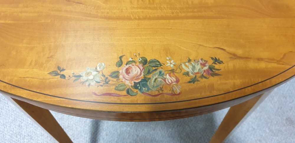 An oval satinwood occasional table featuring painted floral swags on the top surface and ribbon details along square tapered legs. View IV