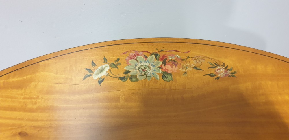 An oval satinwood occasional table featuring painted floral swags on the top surface and ribbon details along square tapered legs. View V