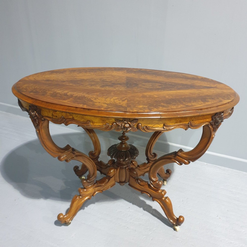 A Victorian oval mahogany centre table featuring a figured veneer top, carved apron with floral motifs, and scrolled legs supported by a central finial and castors.