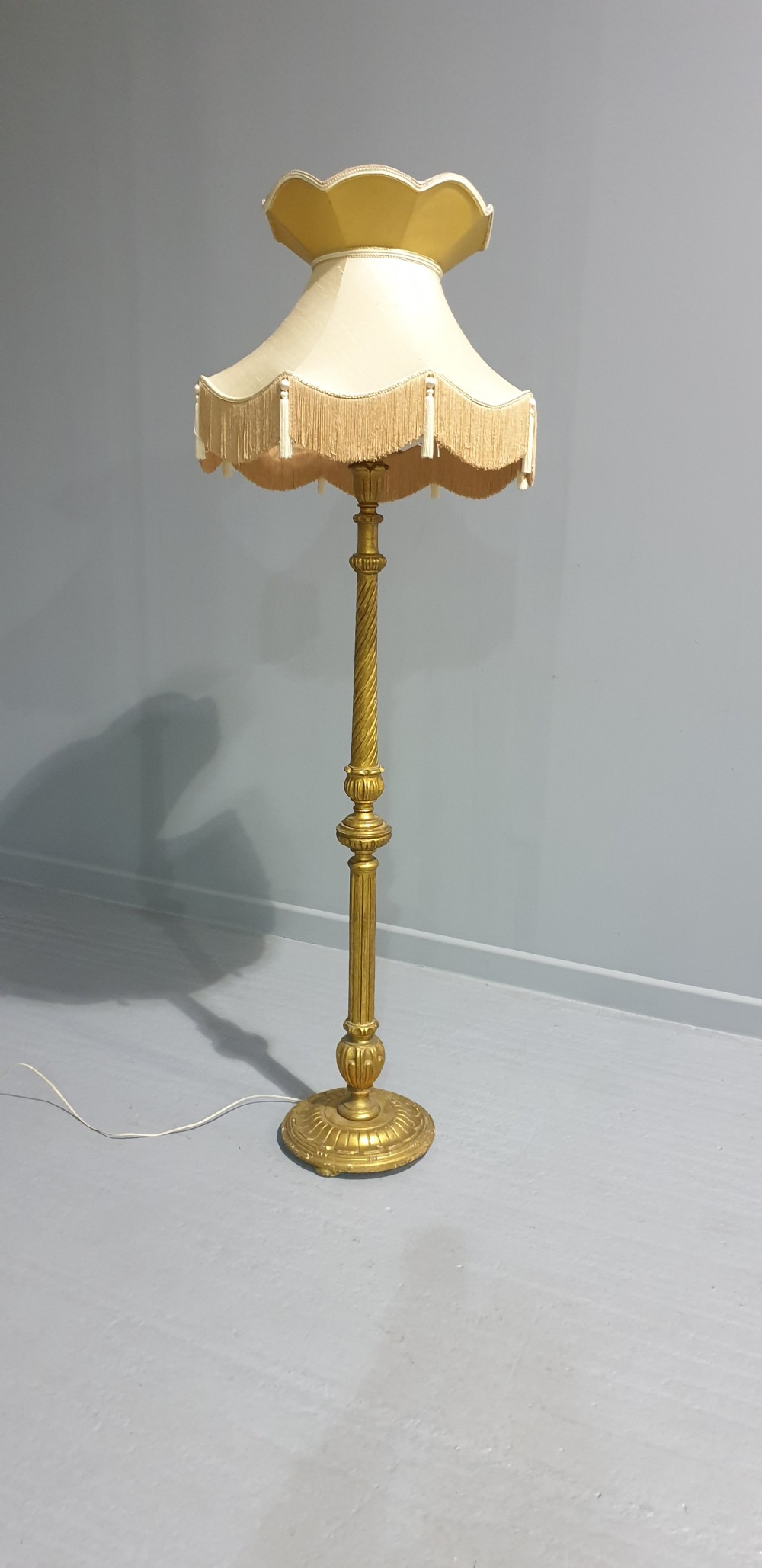 A tall gilded standard lamp featuring a carved fluted column and a cream fabric lampshade trimmed with fringe and tassels.