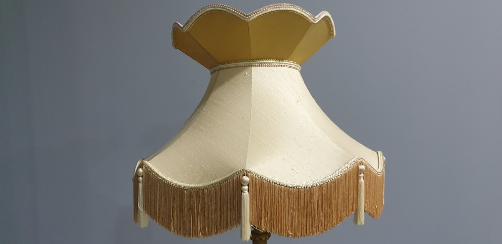 A tall gilded standard lamp featuring a carved fluted column and a cream fabric lampshade trimmed with fringe and tassels. View V