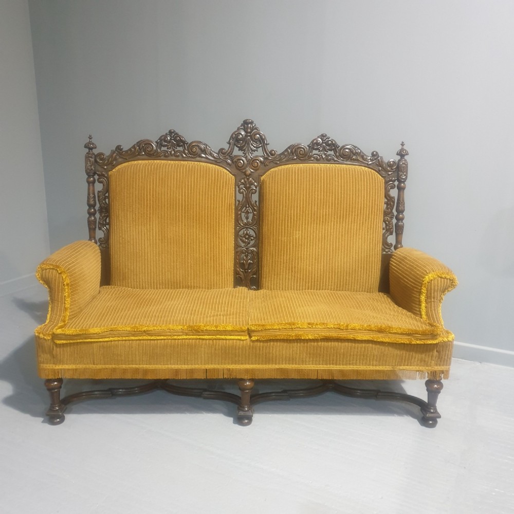 An antique double-backed sofa crafted from dark carved walnut wood with mustard yellow striped upholstery and fringe trim.