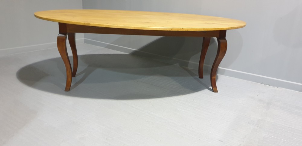 A large French farmhouse dining table featuring a light pine oval top supported by dark fruitwood cabriole legs.