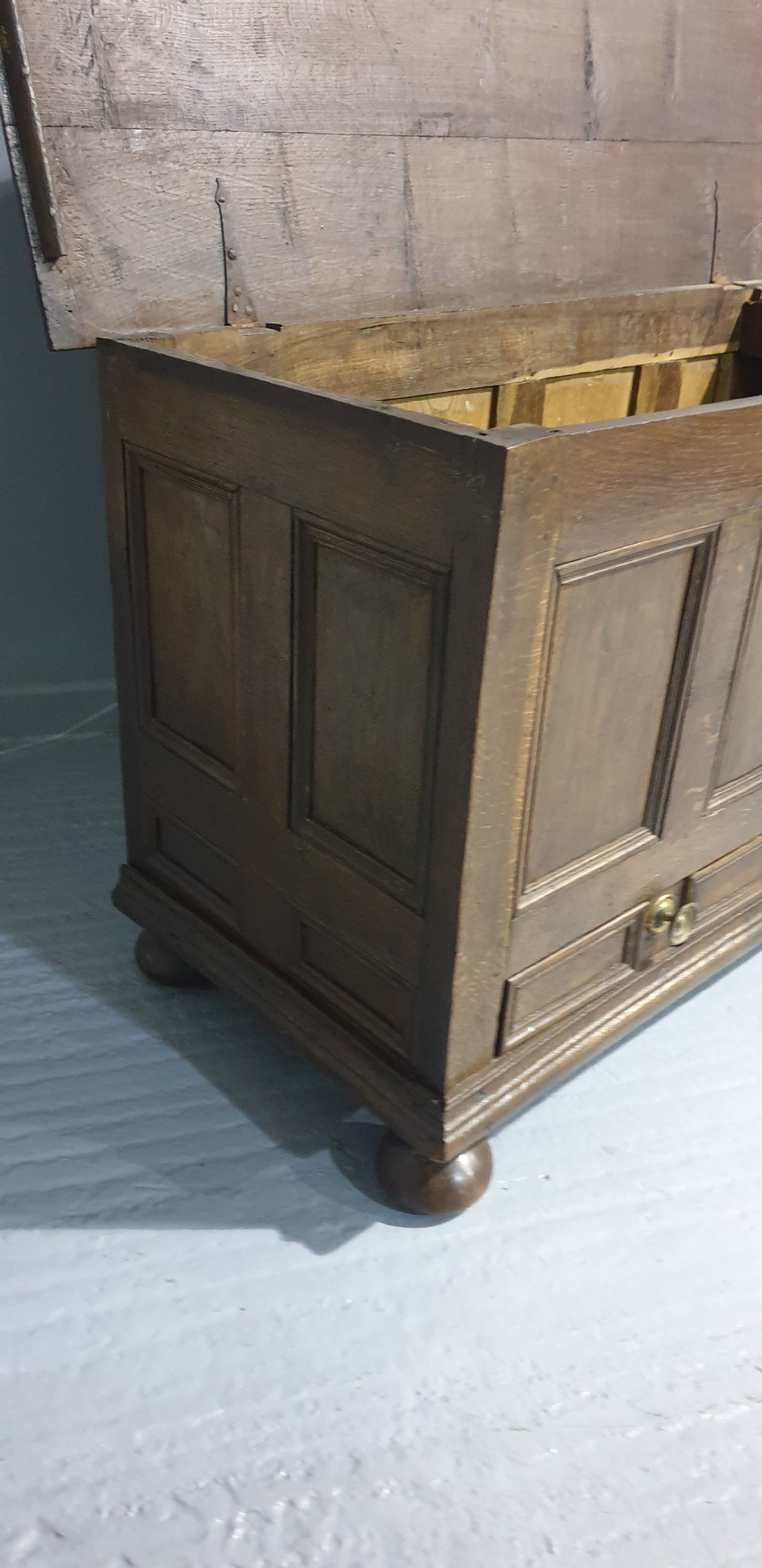 A 17th-century dark oak mule chest featuring four recessed front panels, a bottom drawer with brass knobs, central brass escutcheons, and turned bun feet. View VII