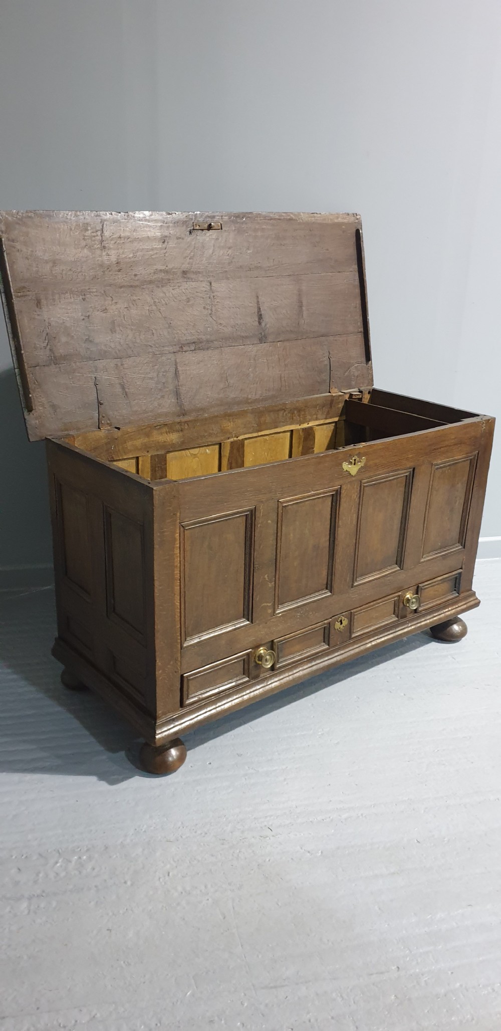 A 17th-century dark oak mule chest featuring four recessed front panels, a bottom drawer with brass knobs, central brass escutcheons, and turned bun feet. View VIII