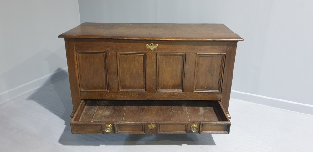 A 17th-century dark oak mule chest featuring four recessed front panels, a bottom drawer with brass knobs, central brass escutcheons, and turned bun feet. View X