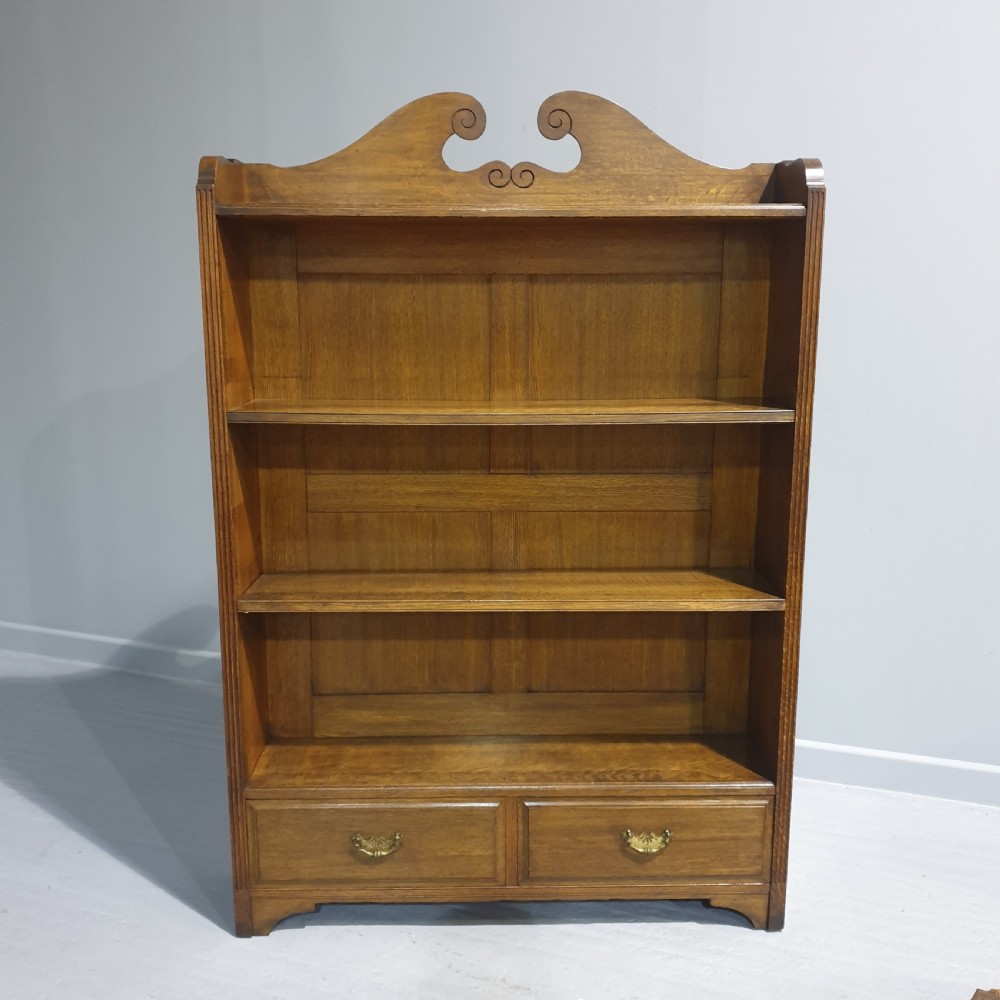 A Victorian medium brown oak open bookcase featuring a carved scroll pediment, adjustable shelves, and two drawers at the base with brass handles.