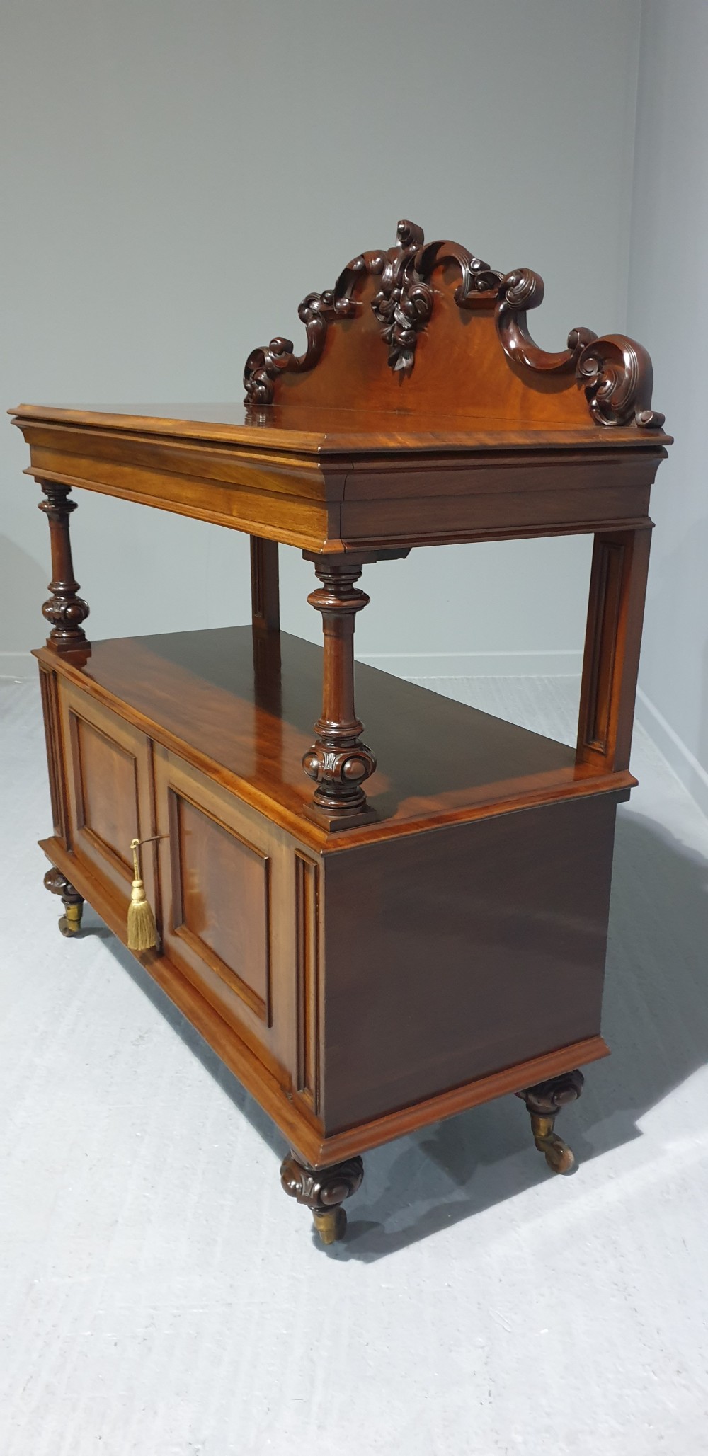 A Victorian mahogany buffet server featuring a carved scroll splashback, turned columns, a two-door lower cupboard, and brass castors. View VIII