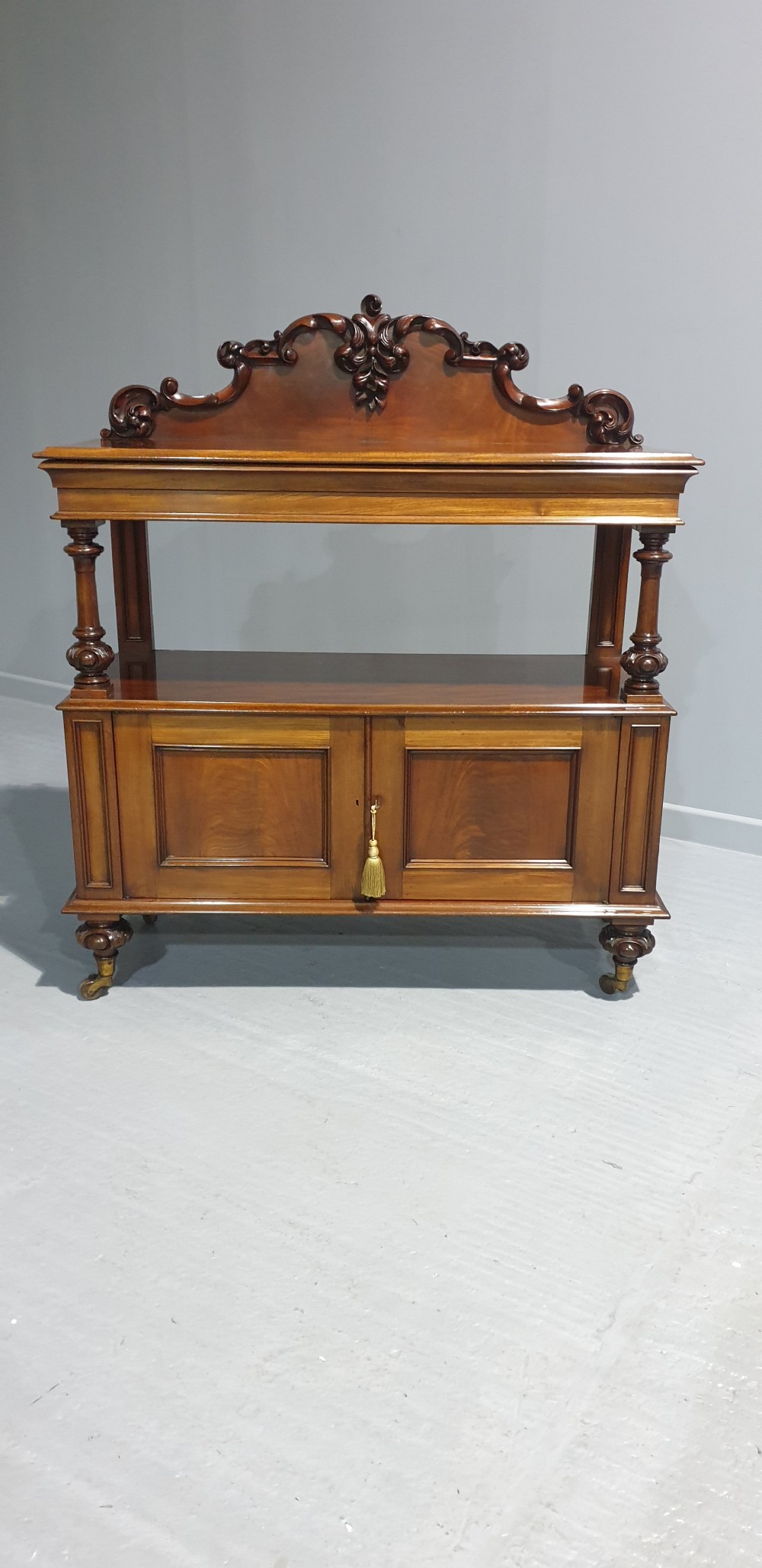 A Victorian mahogany buffet server featuring a carved scroll splashback, turned columns, a two-door lower cupboard, and brass castors. View X