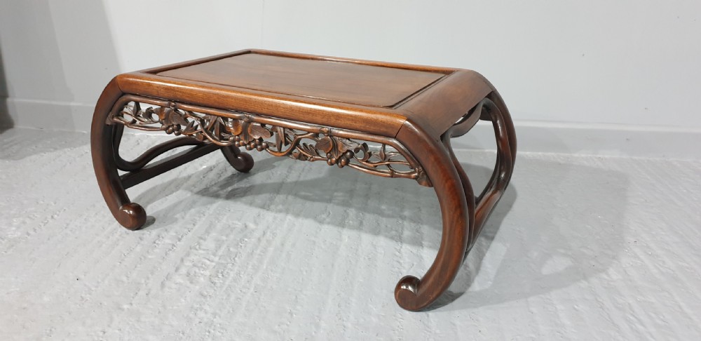 A low warm brown carved hardwood Chinese Kang opium table featuring curved legs and pierced floral carvings along the apron.