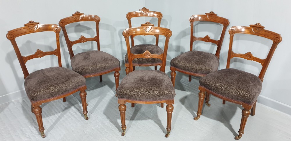 A set of six Victorian carved walnut dining chairs featuring open curved backrests, brown textured fabric seats, and turned front legs fitted with brass castors. View II