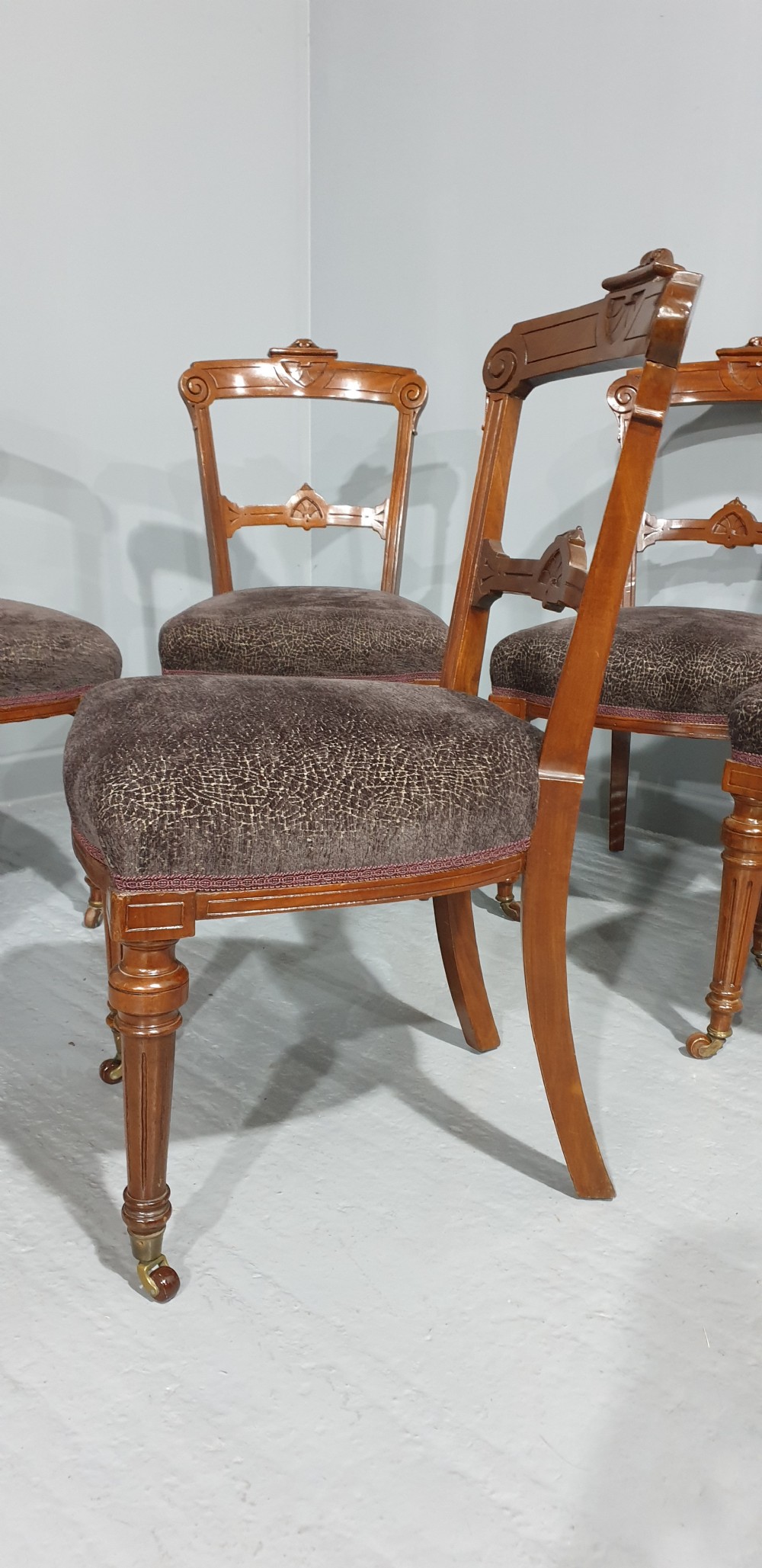 A set of six Victorian carved walnut dining chairs featuring open curved backrests, brown textured fabric seats, and turned front legs fitted with brass castors. View V