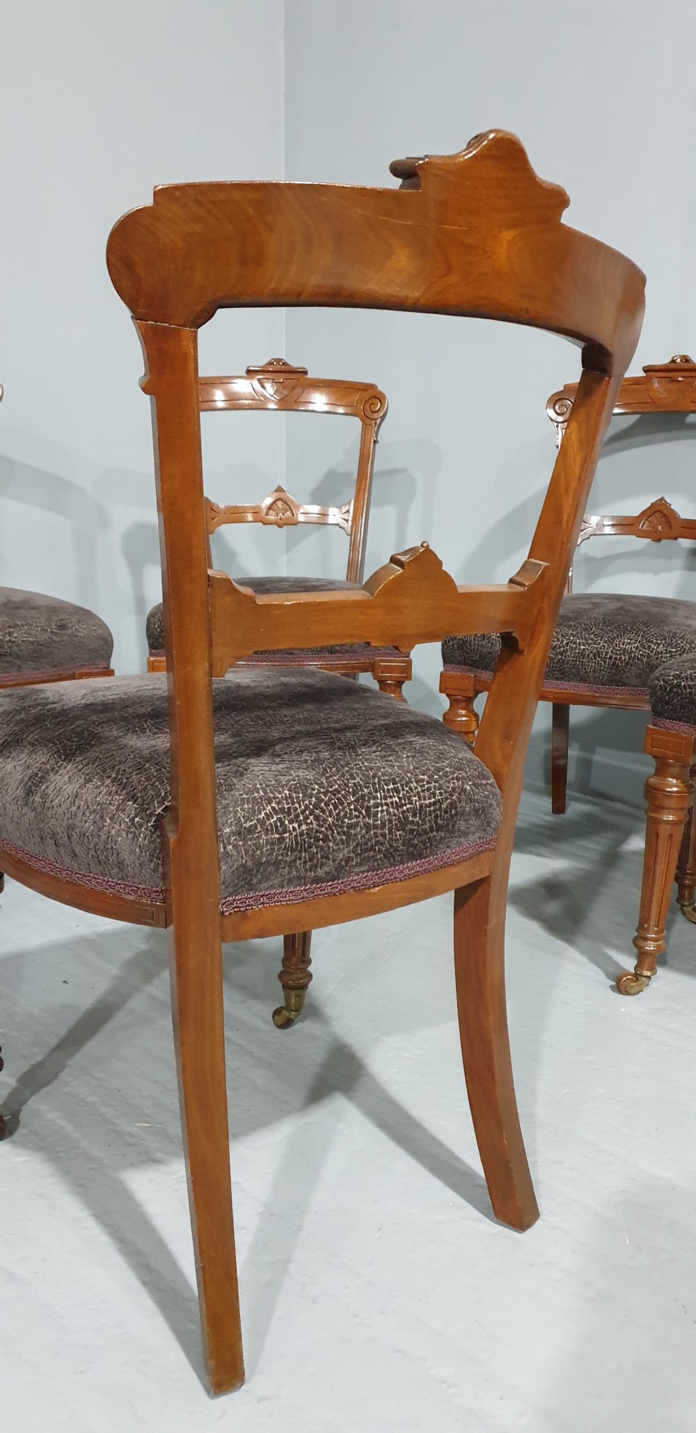 A set of six Victorian carved walnut dining chairs featuring open curved backrests, brown textured fabric seats, and turned front legs fitted with brass castors. View VII