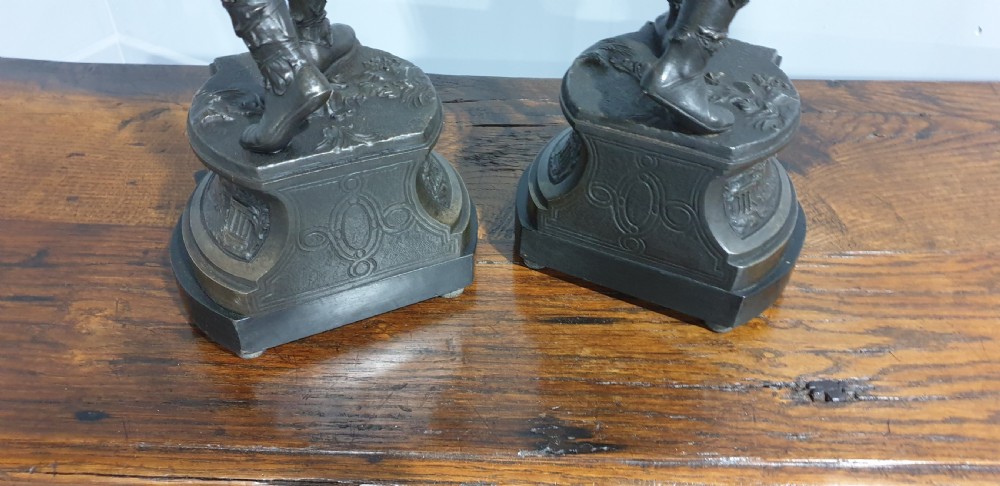 A pair of dark bronze-finished spelter statues depicting classical warriors wearing detailed armour and standing on relief-decorated bases. View X