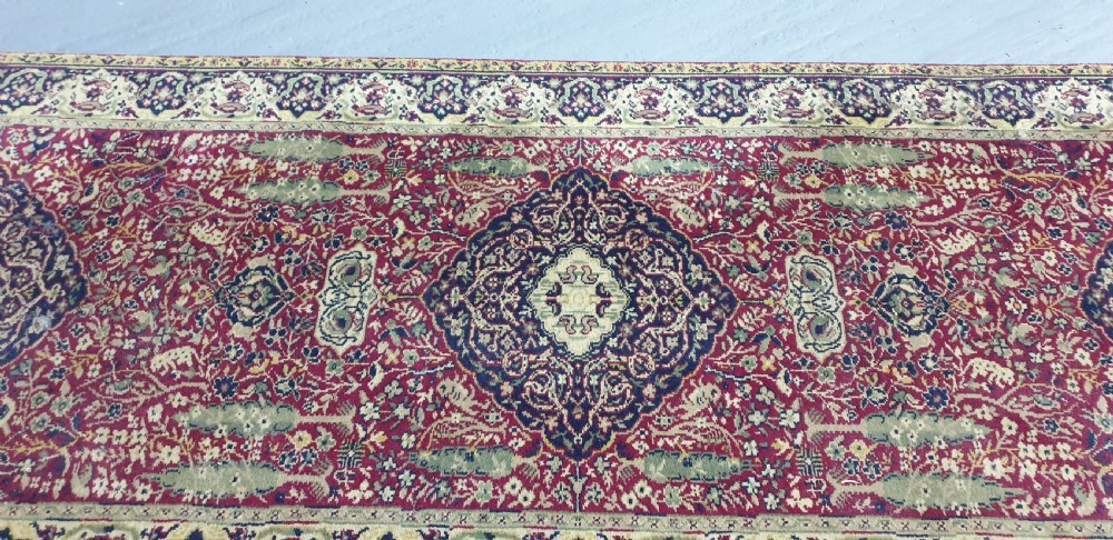 An elongated hand-knotted antique wool runner rug featuring a detailed central medallion and ornate floral motifs in deep red, navy blue, and cream colours. View VI