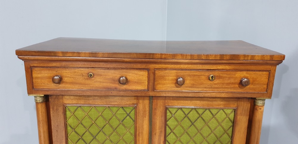 An antique Regency mahogany side cabinet featuring two top drawers over double doors with brass diamond grilles backed by green fabric, flanked by columns. View II