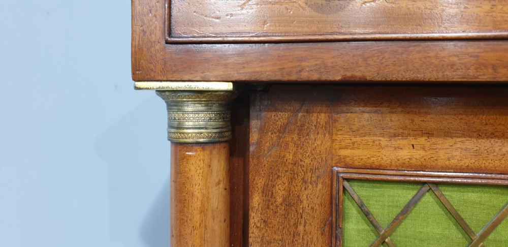 An antique Regency mahogany side cabinet featuring two top drawers over double doors with brass diamond grilles backed by green fabric, flanked by columns. View VIII