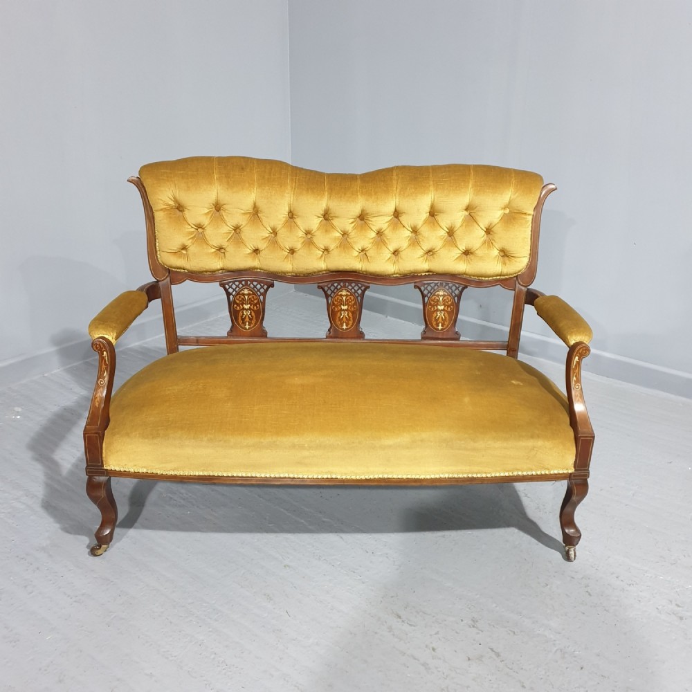 An Edwardian mahogany two-seat settee upholstered in button-tufted gold velvet with decorative marquetry inlays on the backrest and arm supports.