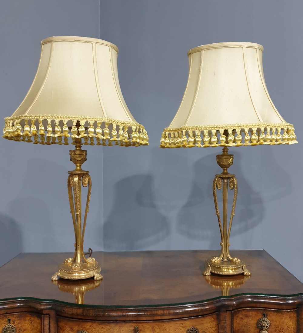 A pair of neoclassical gilded ormolu table lamps with slender cast metal bases and cream fabric shades decorated with tassel fringe.