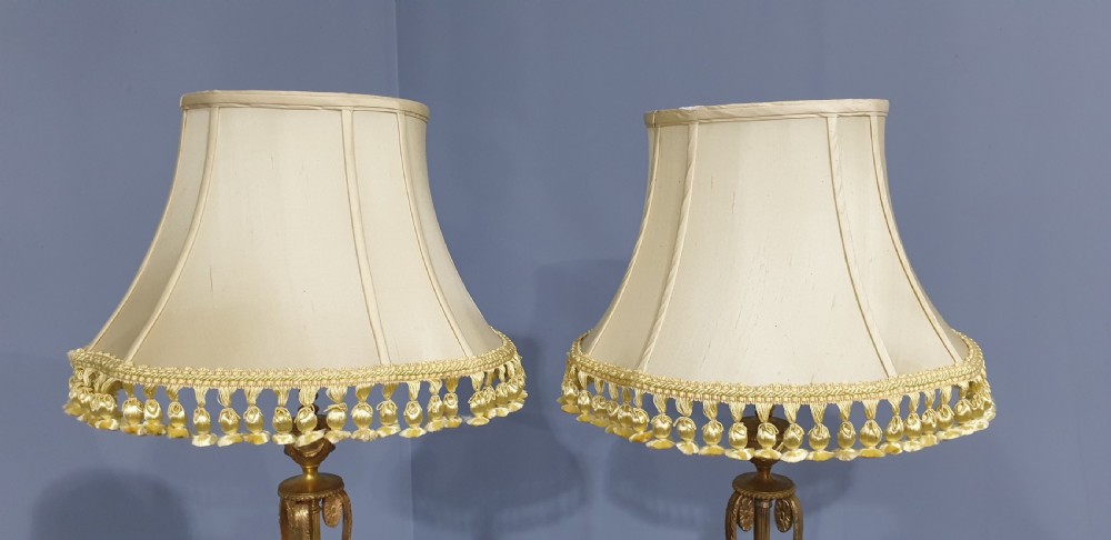 A pair of neoclassical gilded ormolu table lamps with slender cast metal bases and cream fabric shades decorated with tassel fringe. View VIII