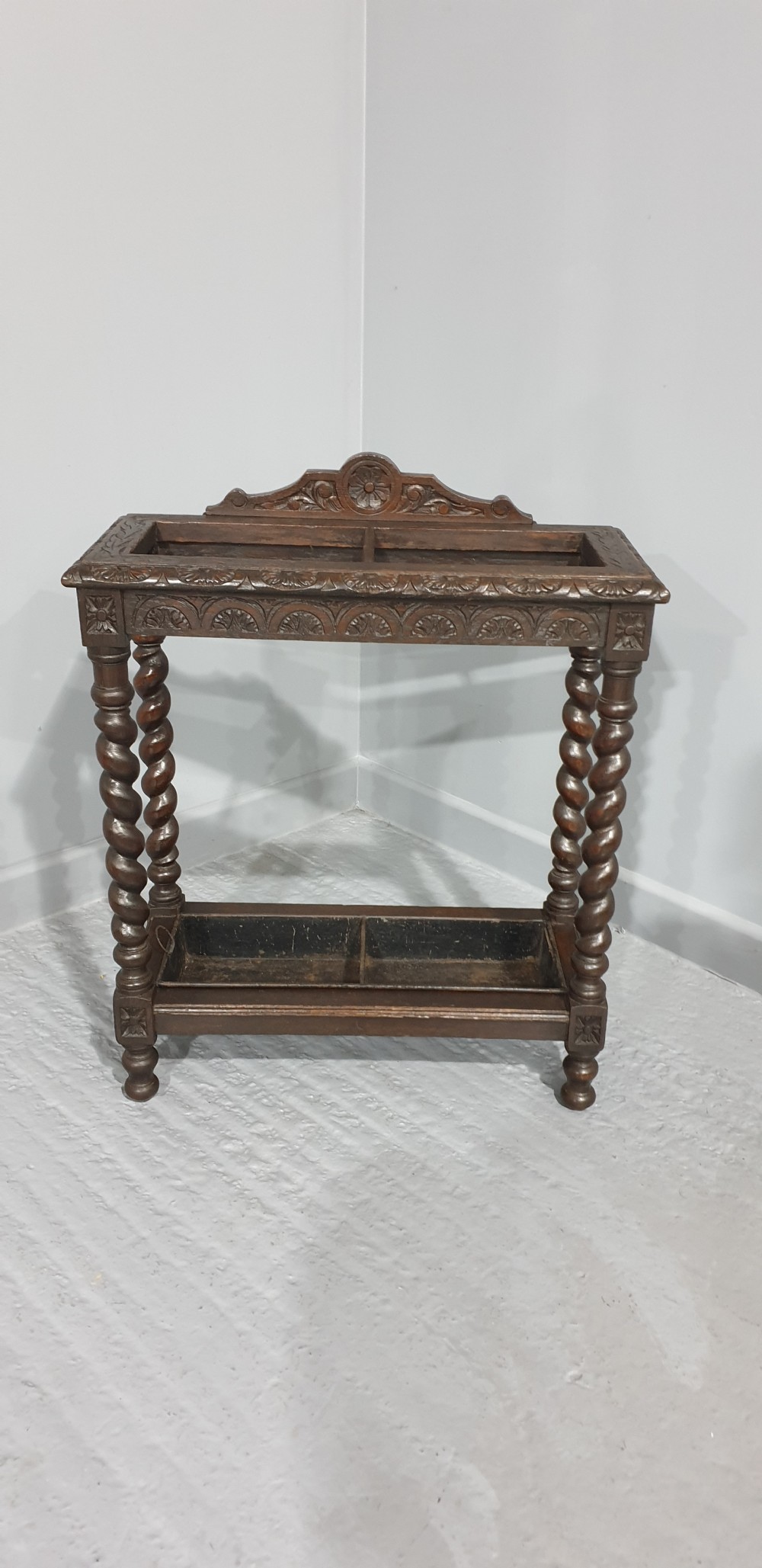 A dark oak stick stand with carved top cresting, barley twist support legs, carved frieze, and lower drip tray.