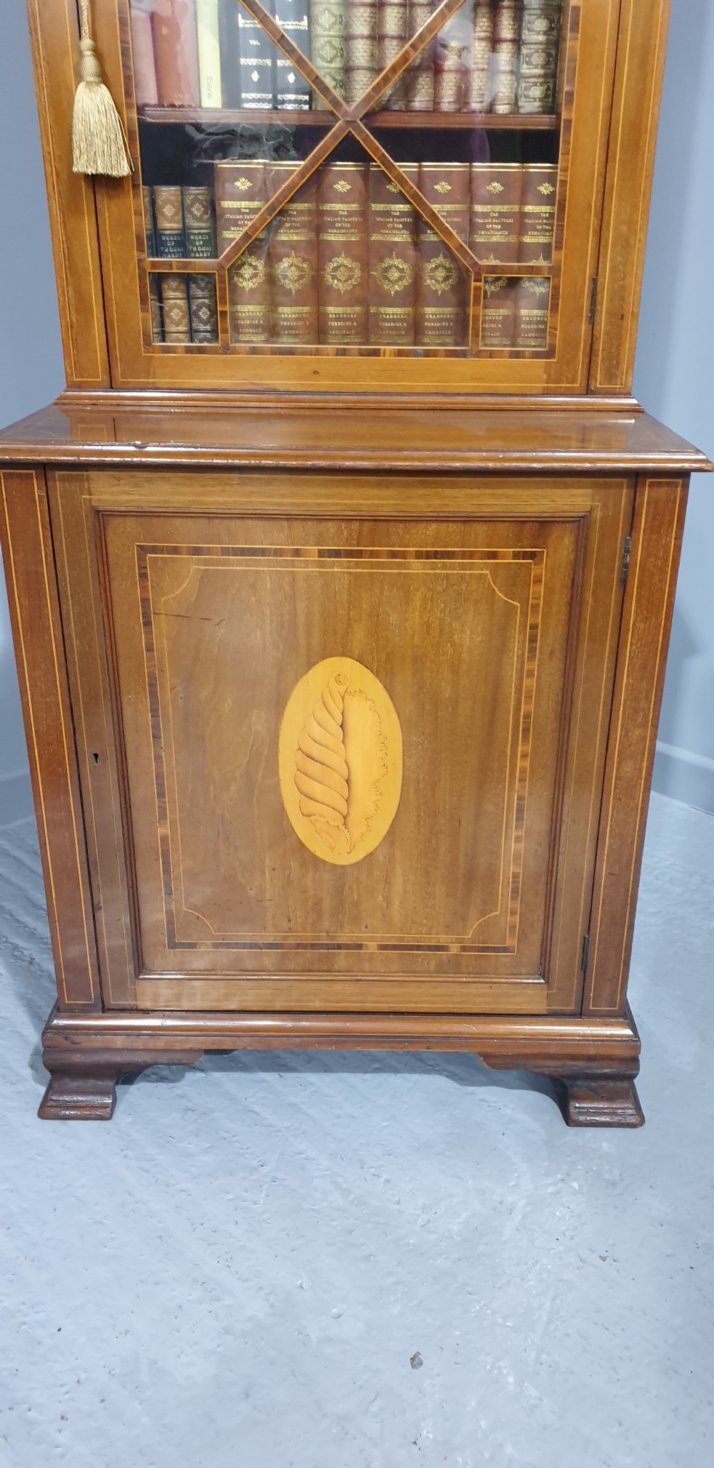 A narrow mahogany standing bookcase featuring a swan-neck pediment top, geometric astragal glazed door, and a lower door with an inlaid oval shell cartouche. View III