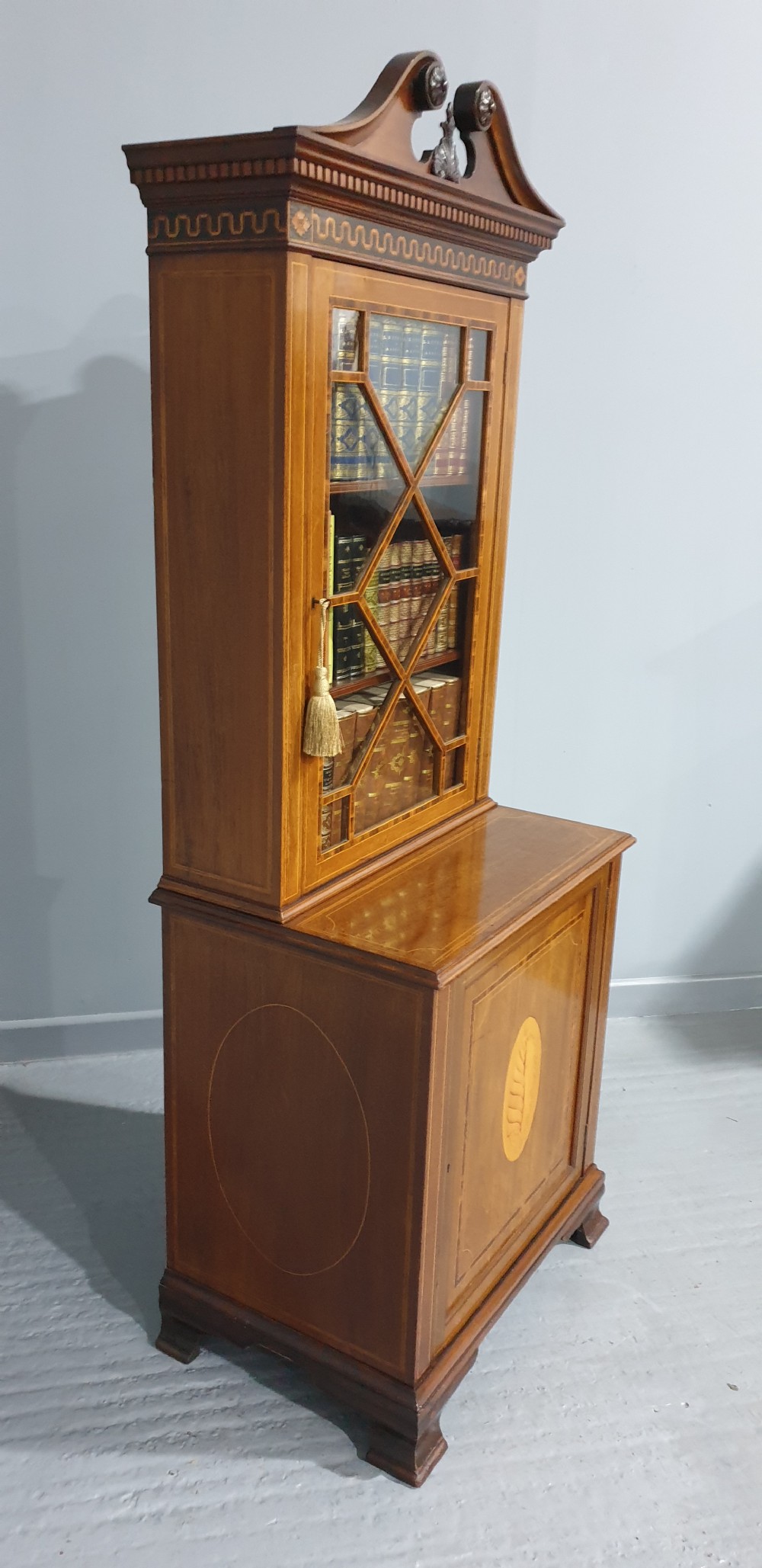 A narrow mahogany standing bookcase featuring a swan-neck pediment top, geometric astragal glazed door, and a lower door with an inlaid oval shell cartouche. View VI