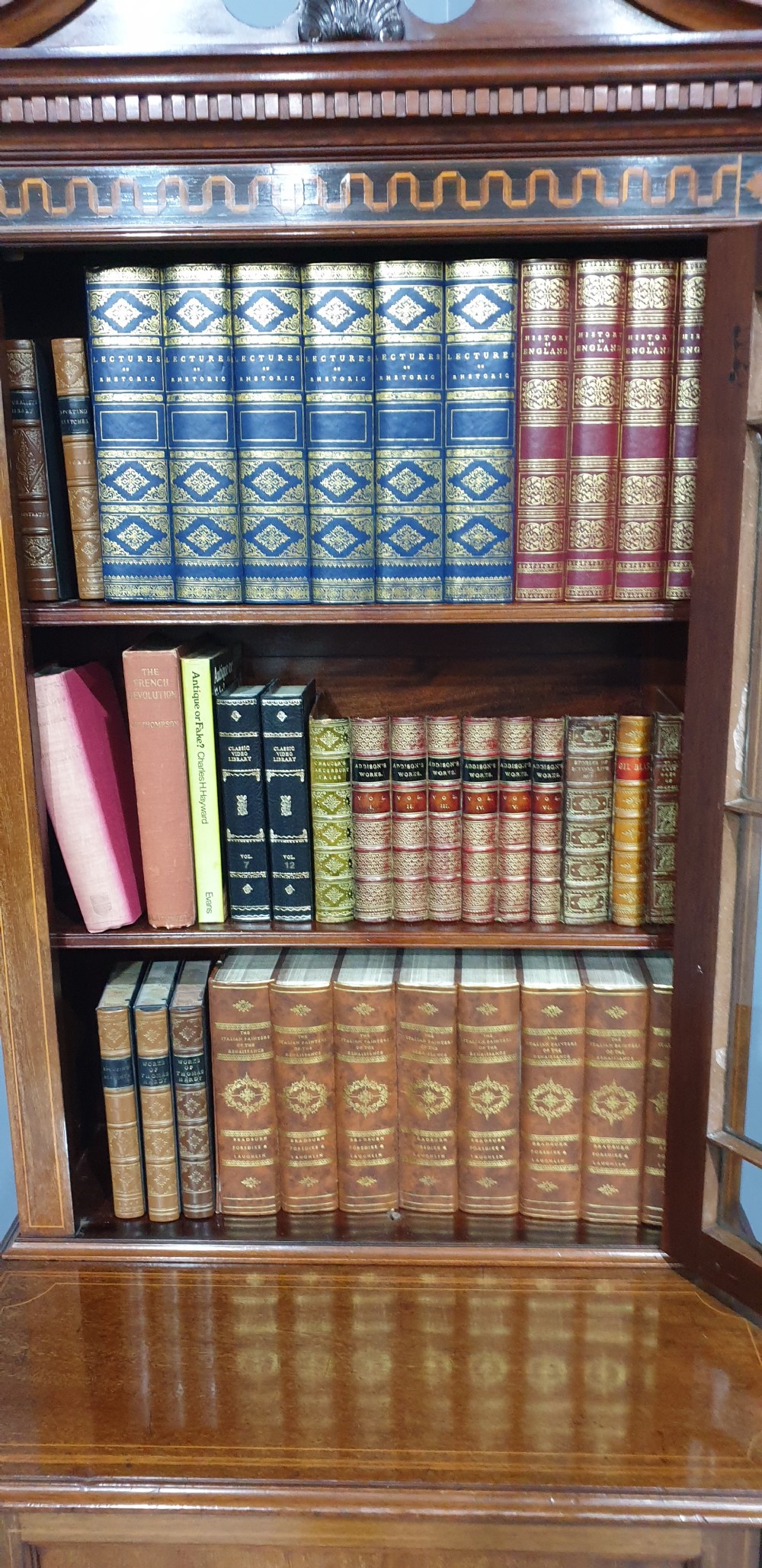 A narrow mahogany standing bookcase featuring a swan-neck pediment top, geometric astragal glazed door, and a lower door with an inlaid oval shell cartouche. View IX