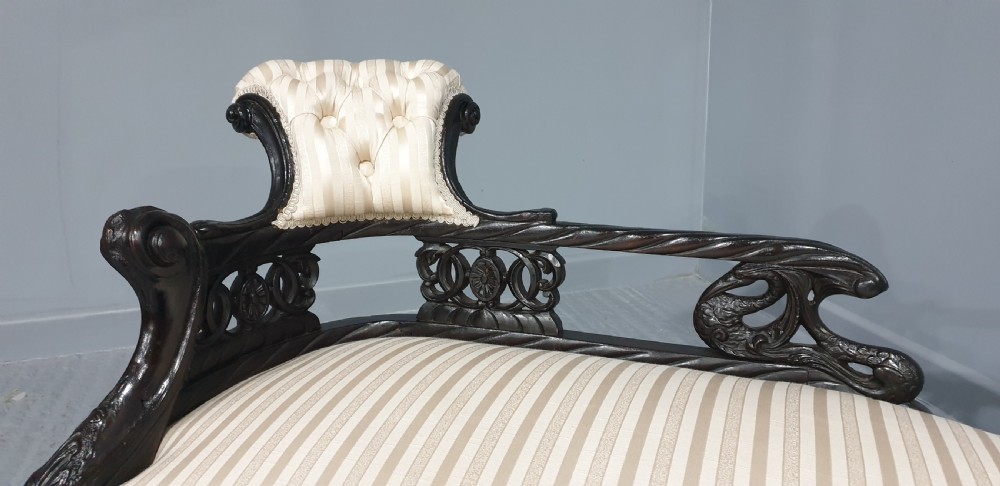 An ebonised wooden miniature chaise longue featuring a button-tufted backrest, cream striped fabric seat, carved frame, and cabriole legs on brass castors. View IV