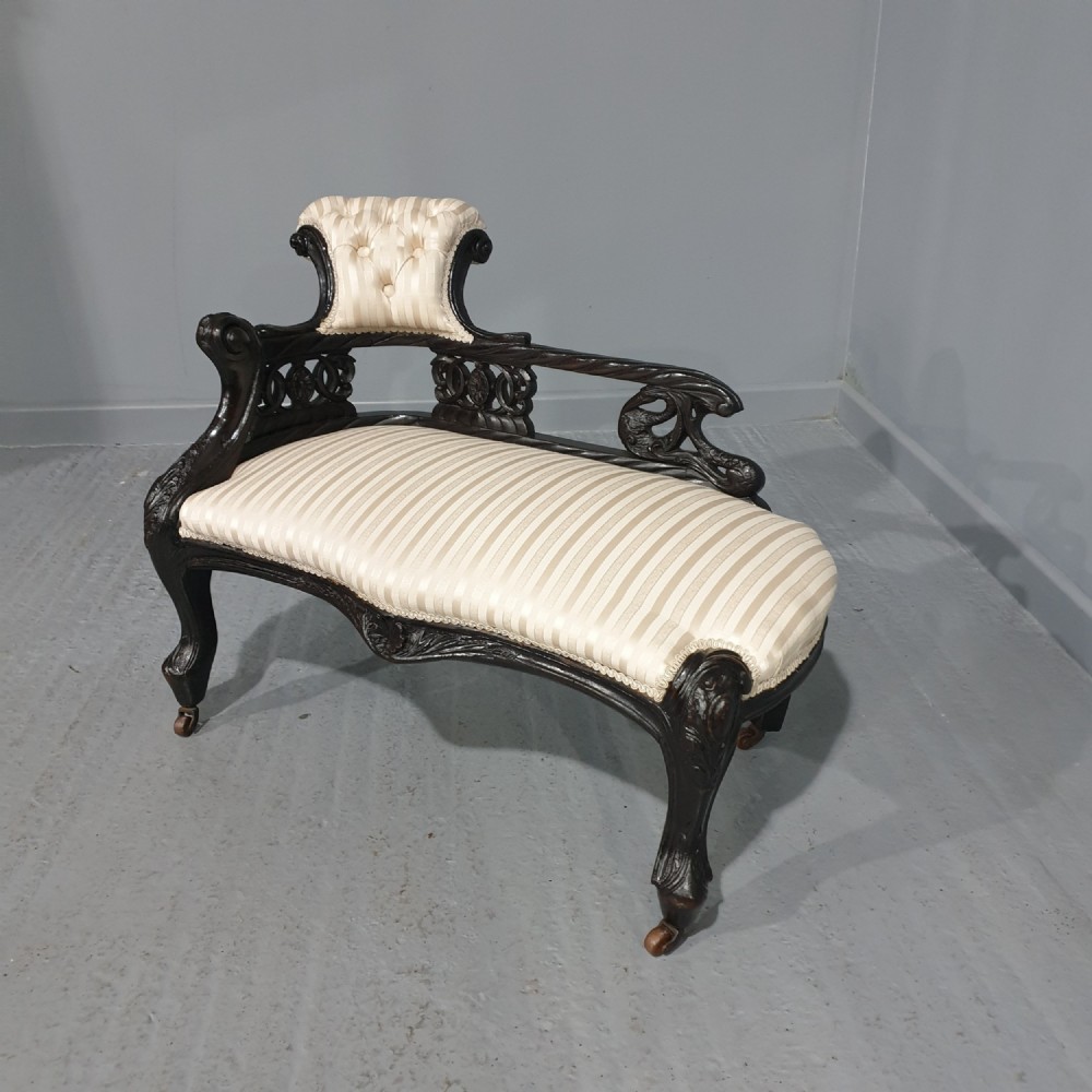 An ebonised wooden miniature chaise longue featuring a button-tufted backrest, cream striped fabric seat, carved frame, and cabriole legs on brass castors.