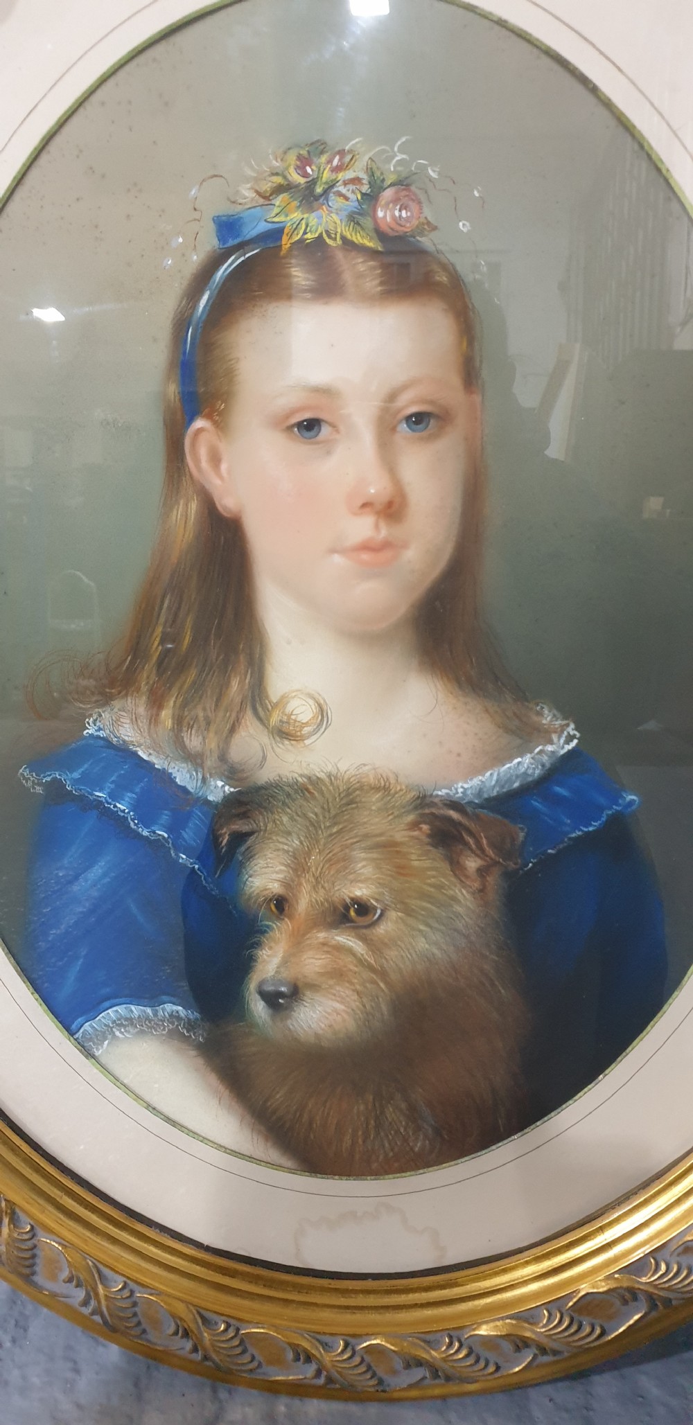 An antique oval Victorian portrait depicting a young girl in a blue dress holding a small dog, set within a carved gold gilded frame. View VI