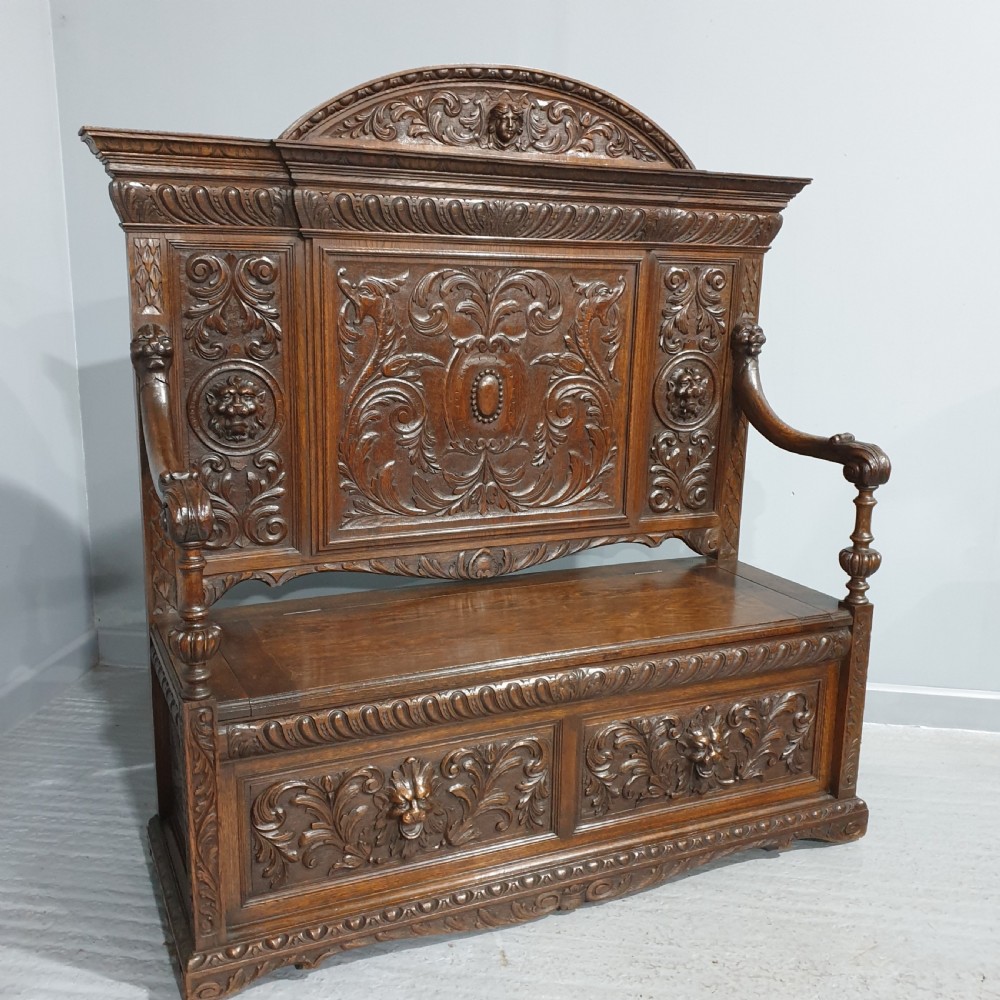 A dark carved oak monks bench with a high arched back, relief foliage and mask carvings, curved armrests, and a hinged seat lid above a box base.