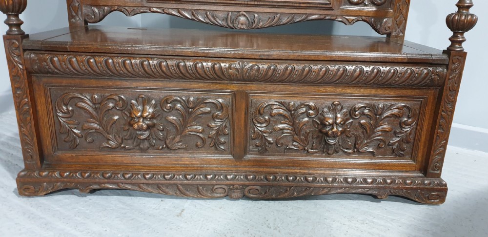 A dark carved oak monks bench with a high arched back, relief foliage and mask carvings, curved armrests, and a hinged seat lid above a box base. View III