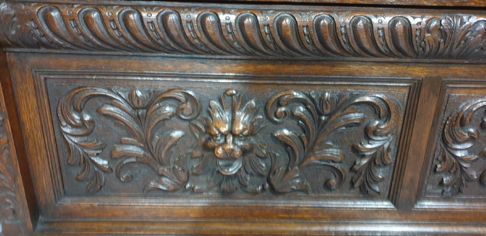 A dark carved oak monks bench with a high arched back, relief foliage and mask carvings, curved armrests, and a hinged seat lid above a box base. View IV