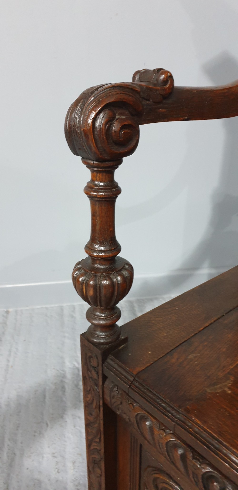 A dark carved oak monks bench with a high arched back, relief foliage and mask carvings, curved armrests, and a hinged seat lid above a box base. View V