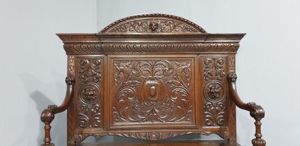 A dark carved oak monks bench with a high arched back, relief foliage and mask carvings, curved armrests, and a hinged seat lid above a box base. View VI
