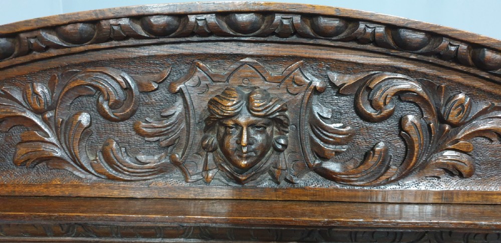 A dark carved oak monks bench with a high arched back, relief foliage and mask carvings, curved armrests, and a hinged seat lid above a box base. View VII