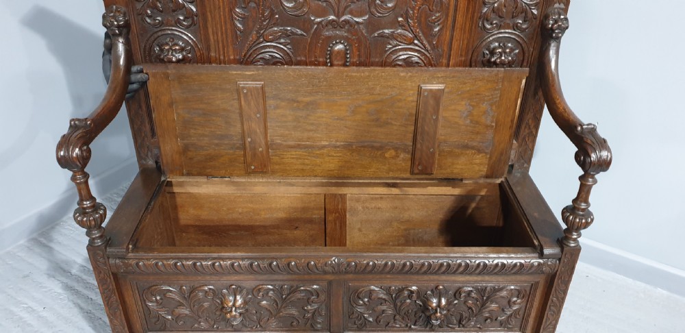 A dark carved oak monks bench with a high arched back, relief foliage and mask carvings, curved armrests, and a hinged seat lid above a box base. View VIII