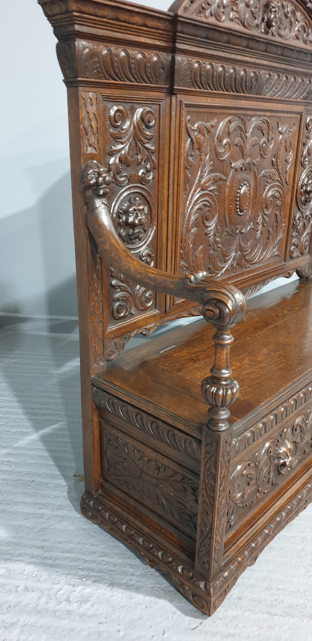 A dark carved oak monks bench with a high arched back, relief foliage and mask carvings, curved armrests, and a hinged seat lid above a box base. View IX