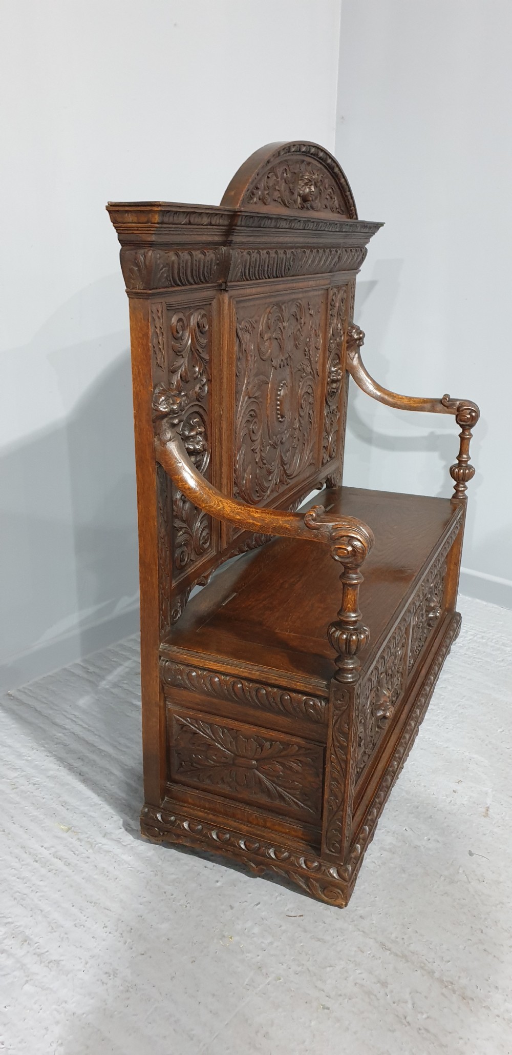 A dark carved oak monks bench with a high arched back, relief foliage and mask carvings, curved armrests, and a hinged seat lid above a box base. View X