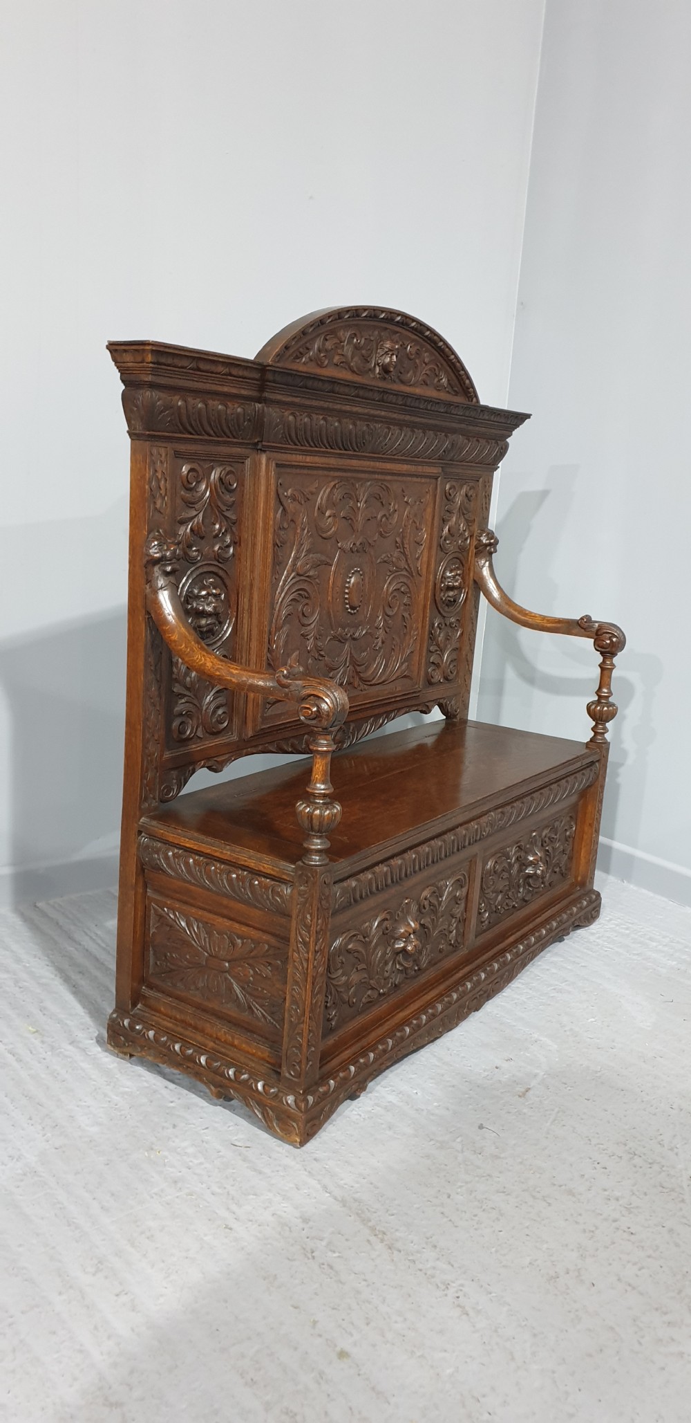 A dark carved oak monks bench with a high arched back, relief foliage and mask carvings, curved armrests, and a hinged seat lid above a box base. View XI