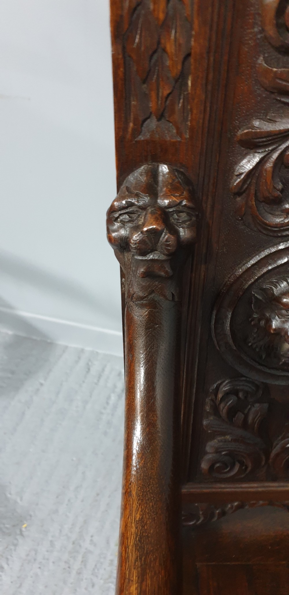 A dark carved oak monks bench with a high arched back, relief foliage and mask carvings, curved armrests, and a hinged seat lid above a box base. View XII