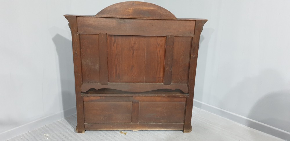 A dark carved oak monks bench with a high arched back, relief foliage and mask carvings, curved armrests, and a hinged seat lid above a box base. View XIII