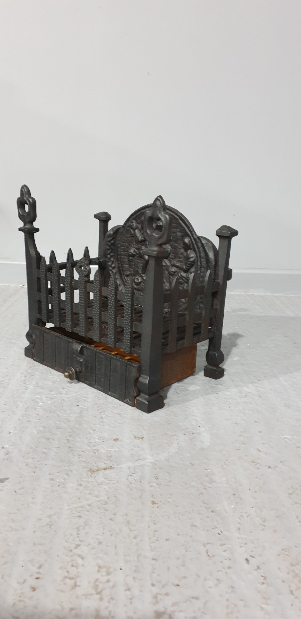 A compact black cast iron fire grate with picket-style front bars, ornamental finials, and a relief-cast backplate depicting figures. View III