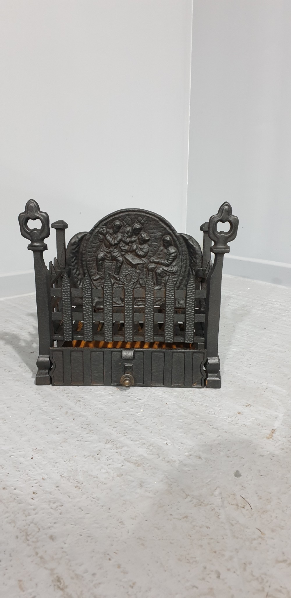 A compact black cast iron fire grate with picket-style front bars, ornamental finials, and a relief-cast backplate depicting figures. View IV