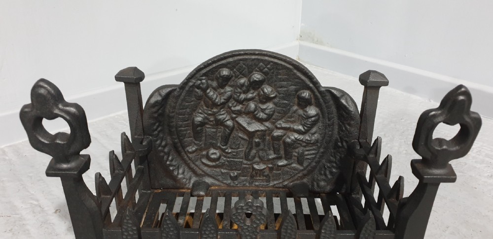 A compact black cast iron fire grate with picket-style front bars, ornamental finials, and a relief-cast backplate depicting figures. View V